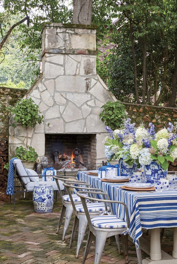 Outdoor table with metal chairs and hydrangea floral arrangements in front of a stone fireplace and Chinese garden stool