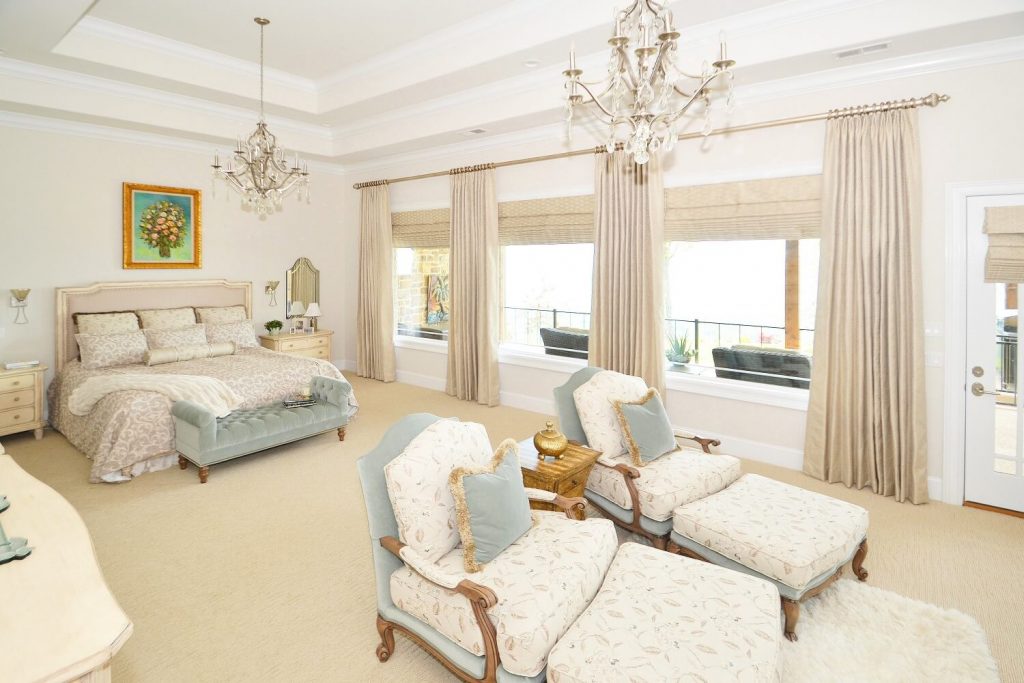 Beige and light blue master bedroom with a bed and two chairs and wall of windows with drapery and roman shades